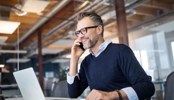 Hymyilevä mies matkapuhelin korvalla katsoo kannettavaa tietokonetta