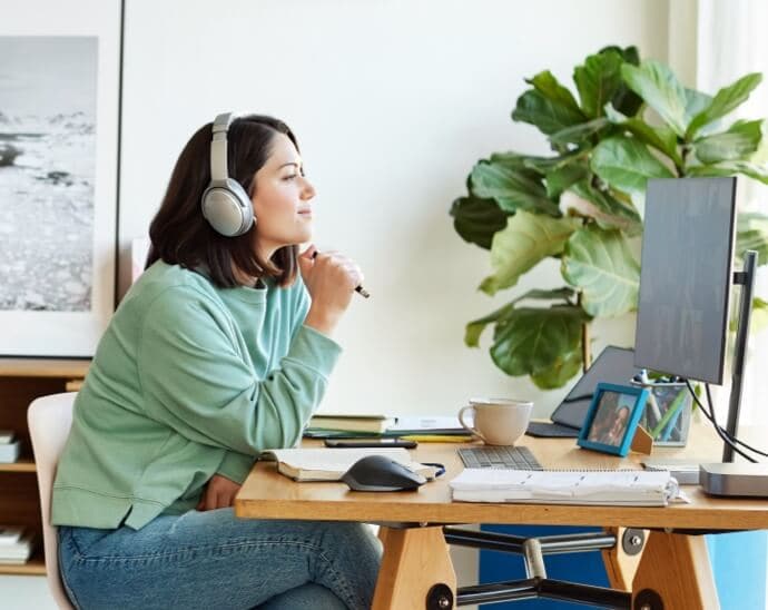 Nainen, jolla on kuulokkeet päässään, pitää kynää kädessään ja katselee tietokoneen näyttöä