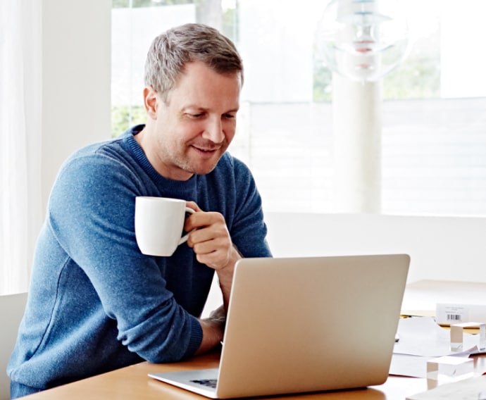Mies käyttää kotona kannettavaa tietokonetta ja juo kahvia.